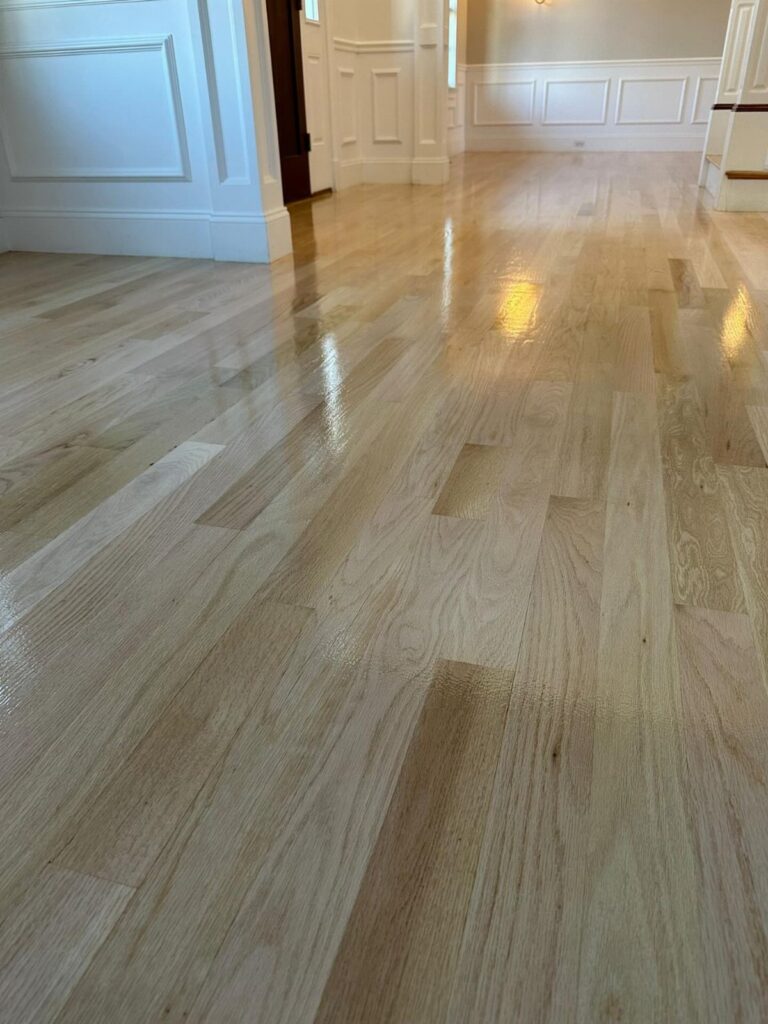Polished light hardwood floor planks with a glossy finish reflecting light in a room with white wainscoting.