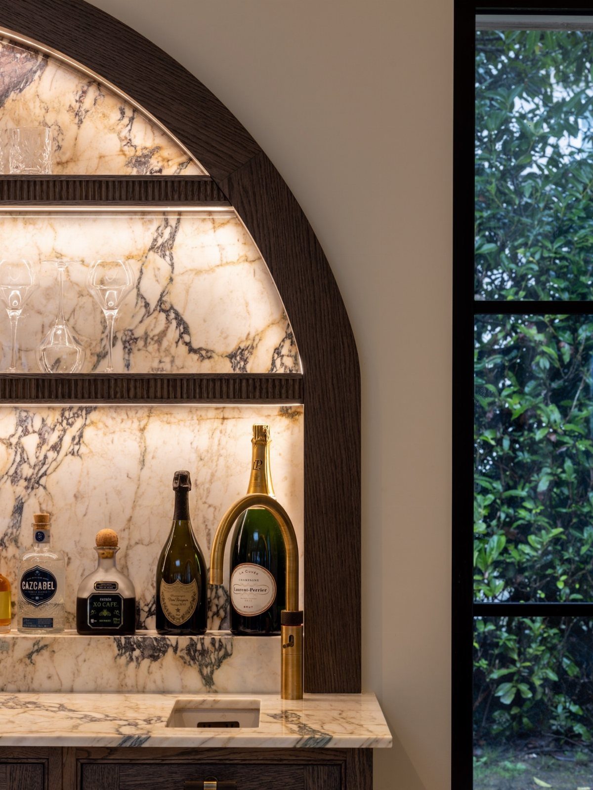 15 Kitchen Renovation Ideas for Getting the Function Back Into the Room 7 A luxurious built-in bar with marble surfaces, gold accents, and various liquor bottles under arched lighting.