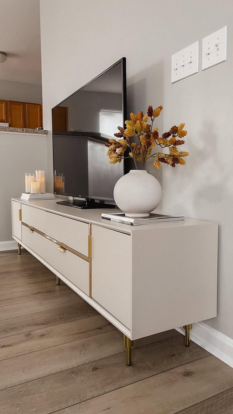 A modern cream sideboard with gold accents holding a television and decorative vase.