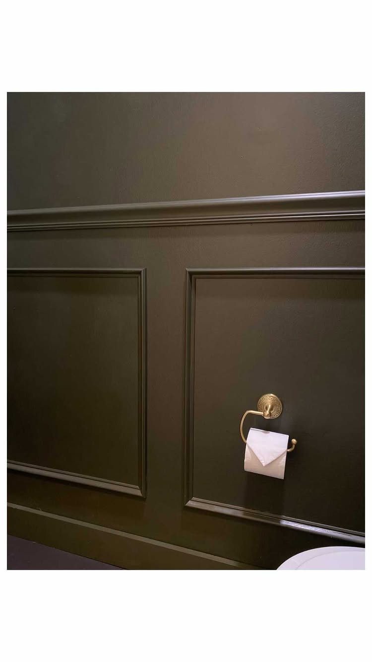 Dark olive green wainscoting with decorative molding and a gold toilet paper holder on a bathroom wall.