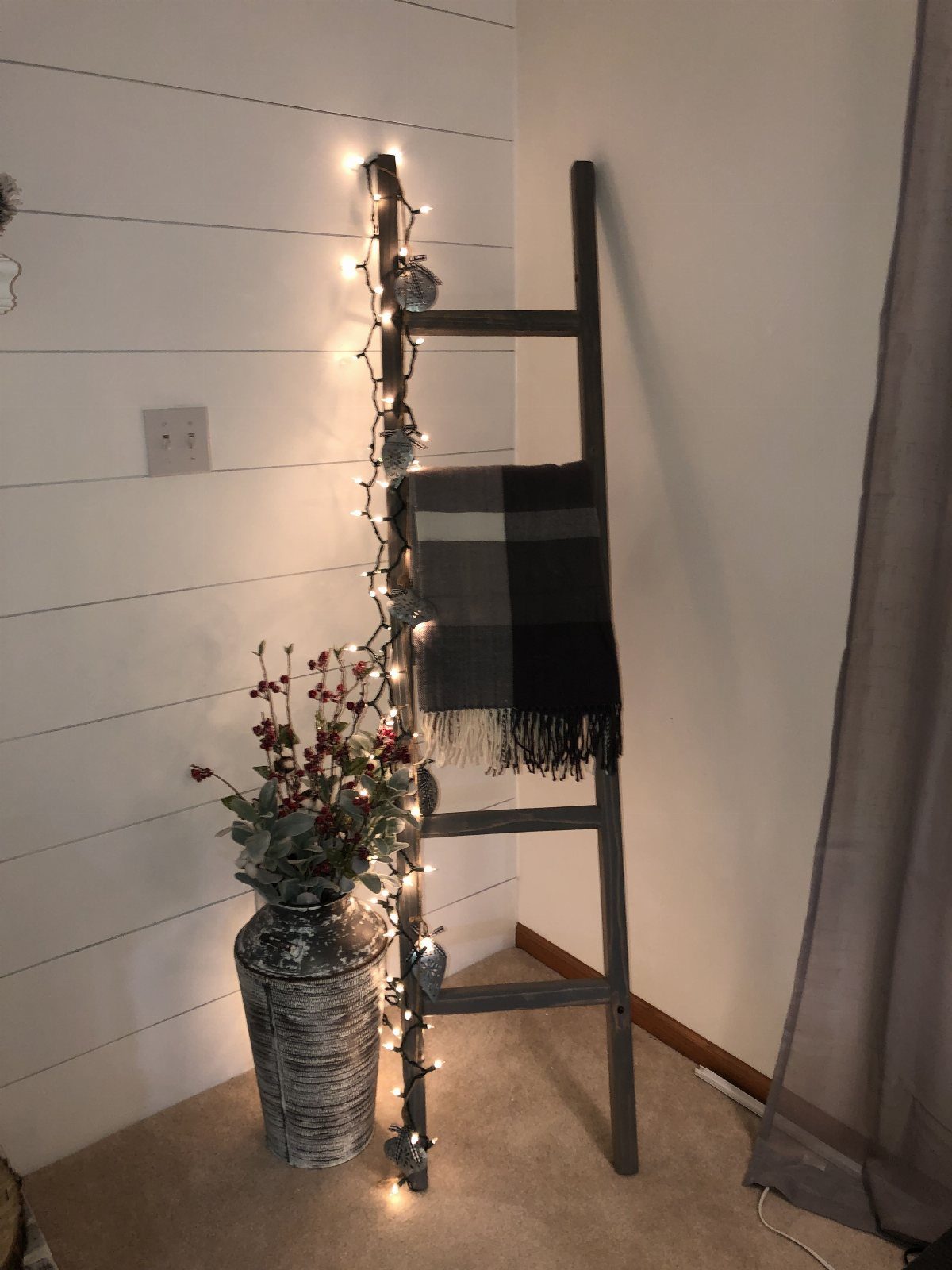 21 Bedroom Wall Decor Ideas With Photos for Every Style 6 A grey wooden decorative ladder with fairy lights and a plaid blanket against a white wall.