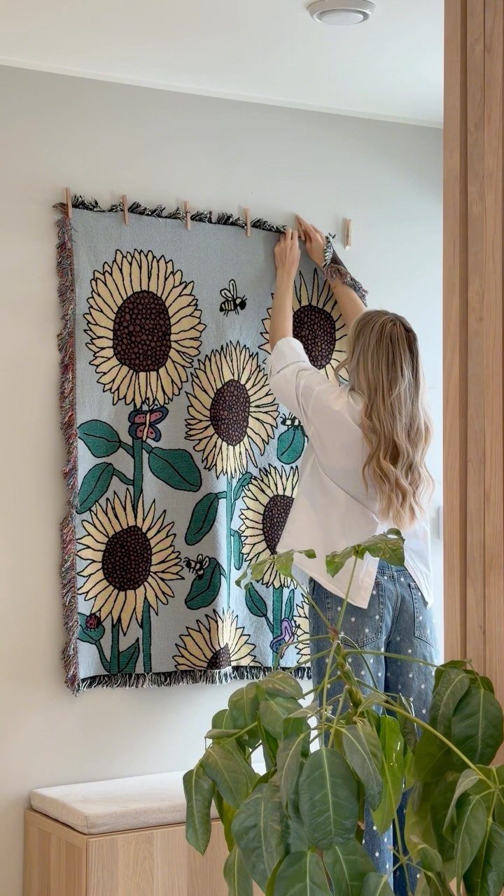 20 Bedroom Wall Designs for Accent, Texture and Art 20 A woman hanging a large sunflower patterned tapestry on a white wall with wooden pegs.