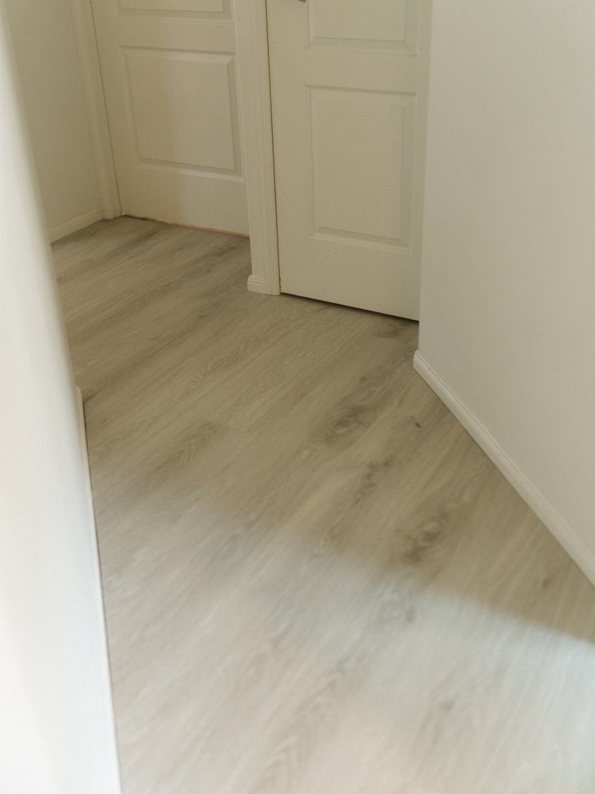 Light greige wood-look vinyl flooring installed in a hallway with white doors and walls.