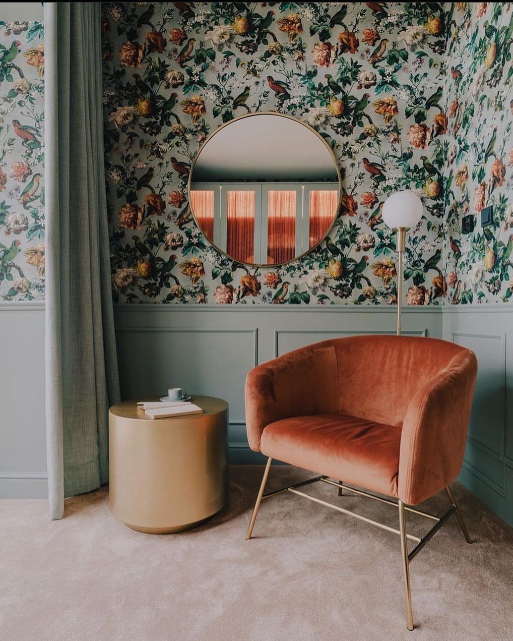Floral wallpapered wall above teal wainscoting with an orange velvet armchair and gold side table.