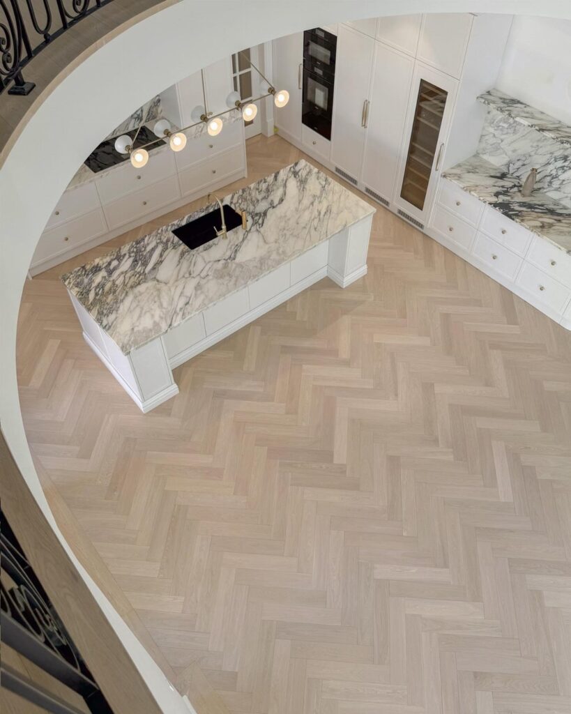 Light wood herringbone pattern flooring in a modern white kitchen with a marble island.