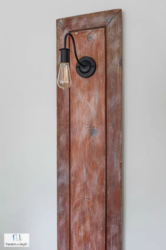 21 Bedroom Wall Decor Ideas With Photos for Every Style 7 A rustic wooden door panel mounted on a wall featuring a black industrial sconce lamp.