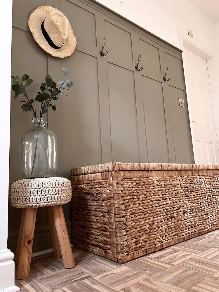 Grey paneled wall with hooks, a straw hat, eucalyptus in a vase, and a wicker chest.
