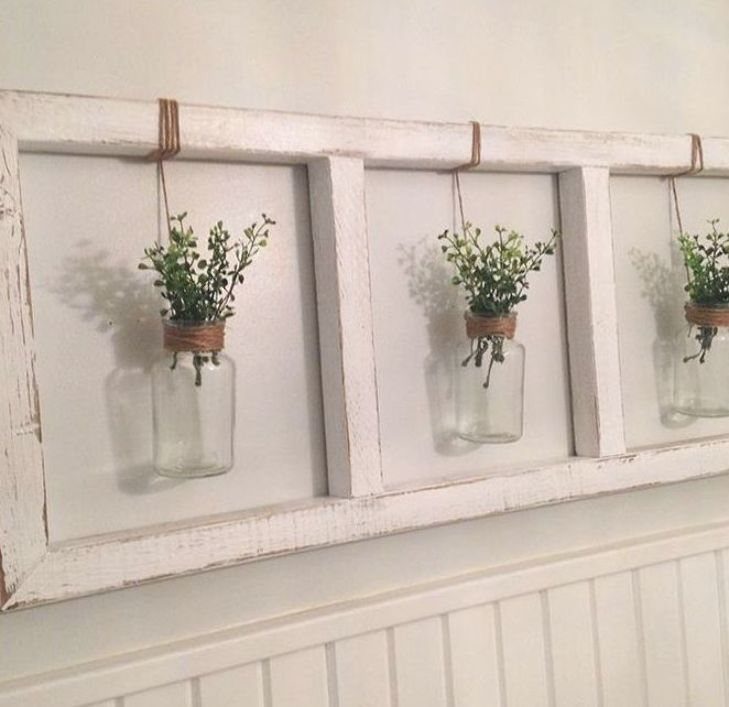 White distressed wooden frame with three hanging glass jars containing green plants on a wall.