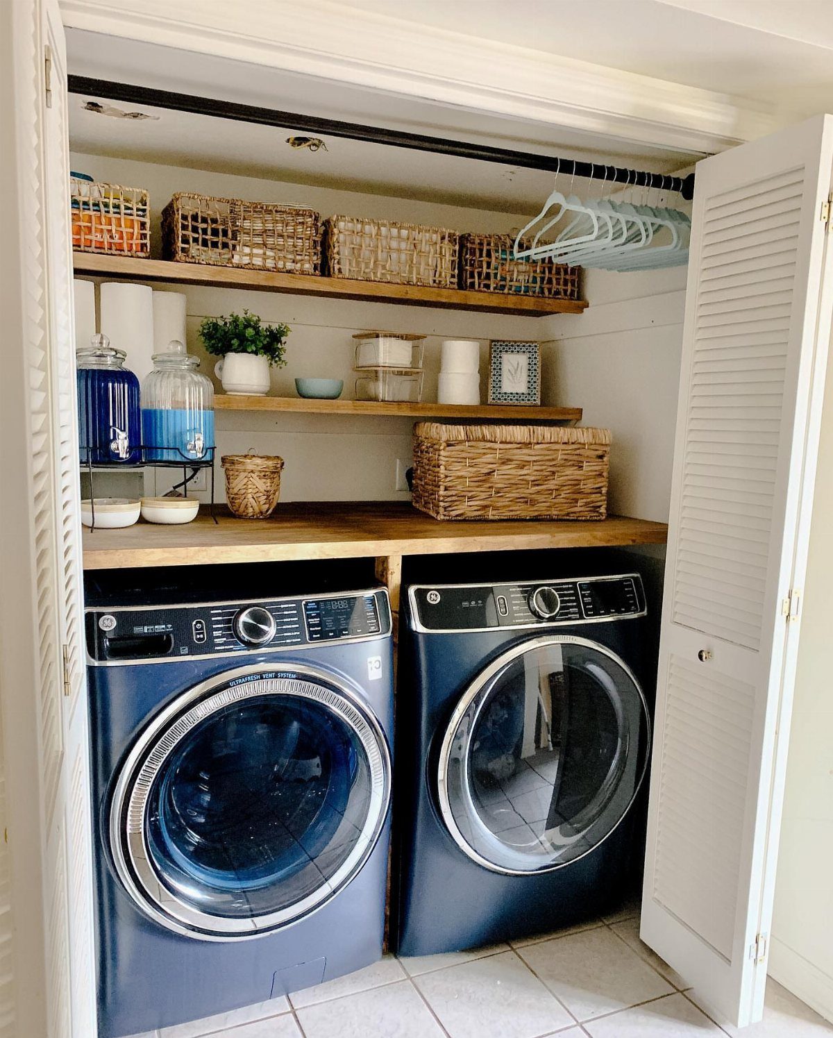 Blue washer and dryer set installed inside a small white louvered laundry closet with wooden shelving.