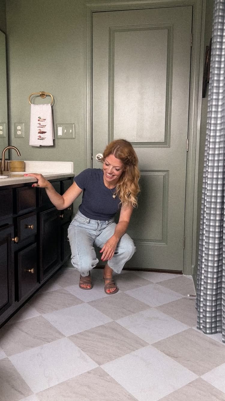 A smiling woman crouches in a bathroom looking at checkered tile-patterned vinyl flooring.