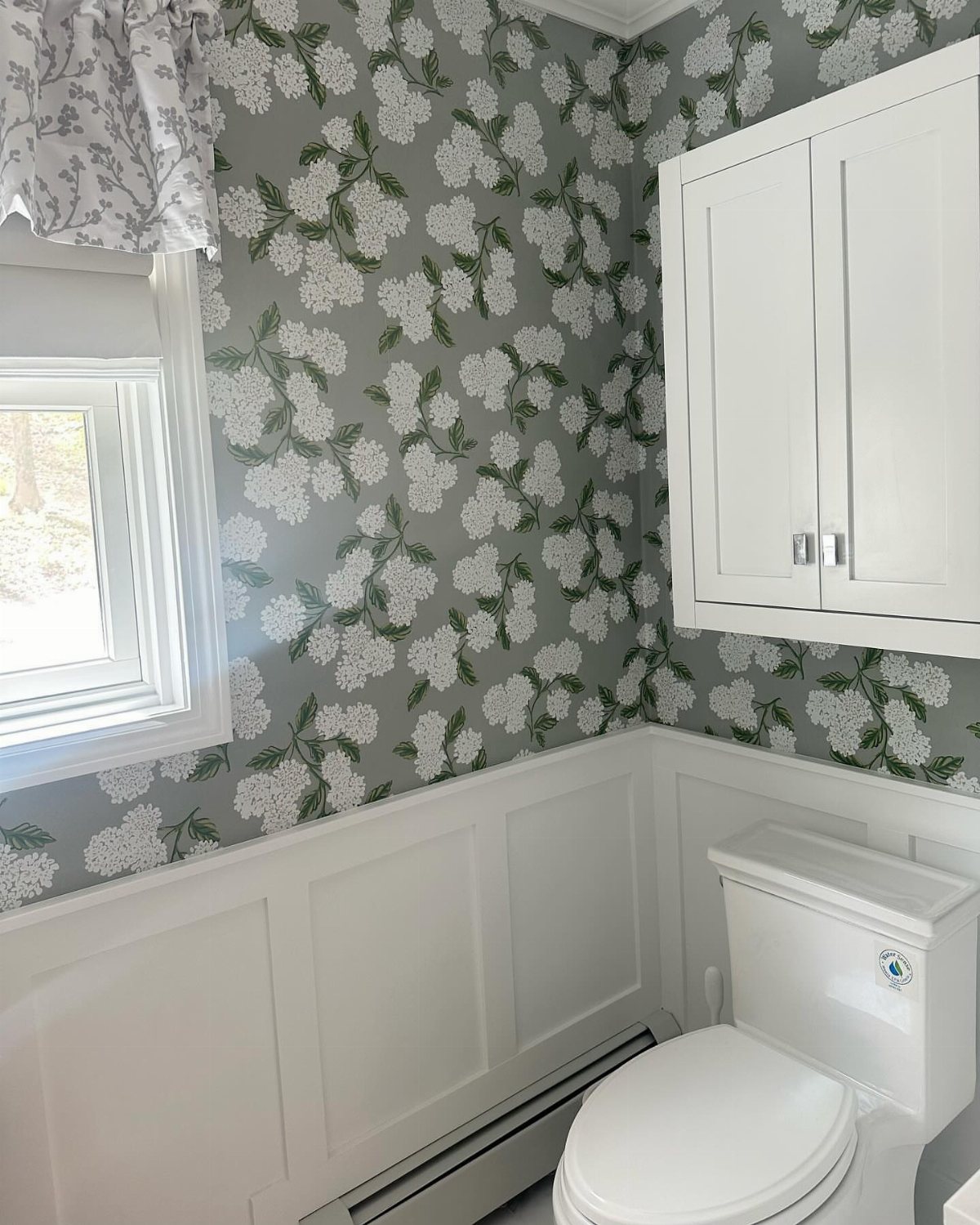 White wainscotting panels installed on a bathroom wall beneath grey floral patterned wallpaper.