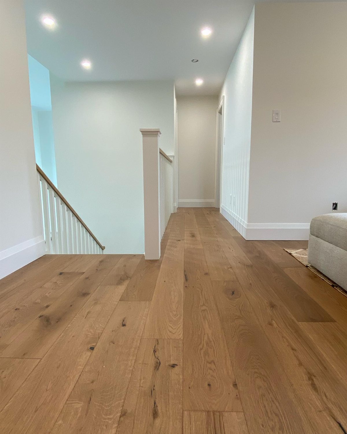 Light brown hardwood flooring planks stretching through a bright hallway next to a white staircase.