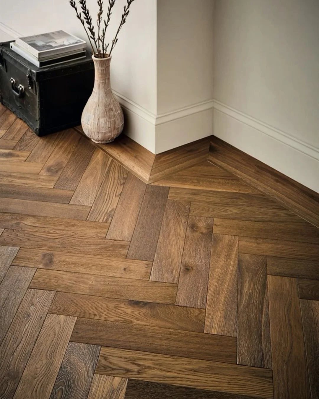 Herringbone pattern warm wood flooring in a room with white walls and a decorative vase.