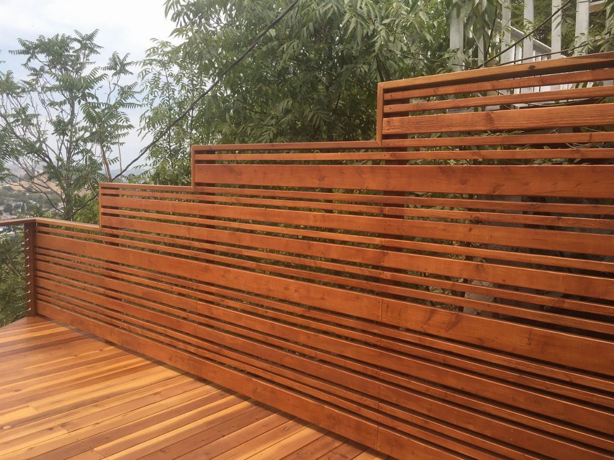 5 Wood Fence Horizontal Ideas for Every Yard 3 A stepped horizontal wood slat fence installed along a wooden deck overlooking green trees.